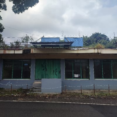 This image depicts a single-story, likely disused commercial building in Cibeureum, Indonesia, viewed from across a paved road under an overcast sky.
The building's facade is predominantly light gray. A central, elevated entryway features closed, green vertically paneled double doors, which exhibit wear and contain faint graffiti. To the left and right of the doors, two large rectangular windows on each side, framed in green, reflect the environment and partially obscure the interior. A concrete step leads up to the central doors. A light brown/beige overhang extends across the facade, above which the flat roof shows signs of disrepair and minor vegetation growth. On the roof, two prominent rectangular blue utility structures are visible, positioned behind a horizontal metallic frame.
In the immediate foreground, a dark, paved asphalt road runs horizontally, featuring a thin white line towards the bottom. Between the road and the building, a narrow strip of unkempt ground with sparse vegetation is present. A low, makeshift barrier comprising thin metal posts connected by white string or tape delineates the property line along this strip.
The background consists of a cloudy, gray sky. Dense green foliage and trees are visible behind and above the building, particularly on the right side. Overhead, thin horizontal lines, possibly power or communication cables, span across the upper portion of the frame. No individuals are present in the scene.