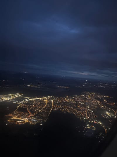 An aerial view of Buštěhrad, Czechia, at night. The scene is dominated by the dark night sky with scattered clouds, and below, the city lights glow with warm orange and yellow hues, creating a network of illuminated streets and buildings. The lights are concentrated in the central and western parts of the image, suggesting a denser urban area. Some areas appear more industrial or commercial, with larger, brighter clusters of lights, possibly indicating infrastructure like airports or industrial parks. The contrast between the dark sky and the illuminated cityscape creates a dramatic visual effect, highlighting the human presence within the nocturnal landscape.