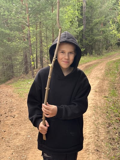 A young child stands on a dirt path in a wooded area, holding a stick horizontally. The child is wearing a black hooded sweatshirt and dark pants. The path runs through a forest of pine trees with sparse green foliage. The ground is covered in dry grass and fallen leaves. This photo was likely taken in Kamennoye, Russia.