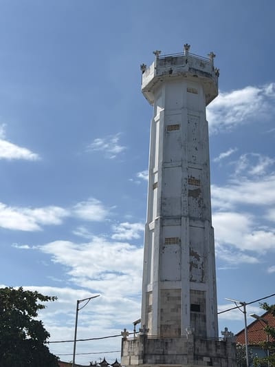 A tall, white, octagonal tower with a weathered facade stands against a clear blue sky with scattered white clouds. The tower has a decorative crenellated top and several small, ornate windows along its height. At the base of the tower, a stone-like platform is visible, also with decorative elements. In the foreground, to the left, a portion of a lush green tree is visible. To the right of the tower, a portion of a red-tiled roof and a modern streetlight with a solar panel are present. The scene suggests an outdoor location in a place with a bright, sunny climate. The description aligns with the general appearance of the landmark known as the Water Tower of Kampung Toyapakeh in Bali, Indonesia, a structure built to store and distribute water.