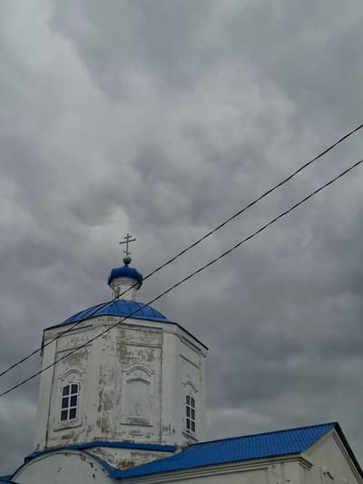 An outdoor, low-angle shot captures a weathered white church building set against a uniformly grey, cloudy sky. The church features a prominent blue dome, topped with a small, dark cross, and portions of its roof are also a vibrant blue. The white plaster on the church walls shows significant signs of age, with peeling paint and visible textures of decay. Rectangular windows with white frames are visible on the church's tower, alongside arched decorative elements carved into the facade.

Three dark power lines stretch diagonally across the upper portion of the frame, partially obscuring the church structure and the overcast sky. The sky is a muted grey, indicating a heavily cloudy day with no direct sunlight. The scene is static and devoid of any human activity. The architectural style, particularly the dome and cross, suggests an Eastern Orthodox church, aligning with the provided location context of Yablonovo, Russia. There is no visible text in the image.