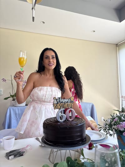 In this image, a woman with long dark hair, wearing a strapless pink and white floral dress, smiles as she holds a glass of orange juice. She is celebrating a birthday, indicated by a "Happy Birthday" cake topper with the number "60" visible. To her left, a large chocolate cake sits on a cake stand. Behind her, another person, likely a child, with long dark hair is partially visible, wearing a pink and white floral outfit. The setting appears to be indoors, possibly a home or event space, with a light-colored wall and a table covered in a light blue cloth. A bouquet of pink and white flowers adds a festive touch. The lighting suggests it is daytime.