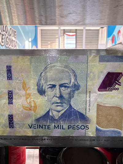 A close-up shot shows a 20,000 Colombian peso banknote. The bill features a portrait of Juan Bautista Alberdi and the phrase "VEINTE MIL PESOS" (twenty thousand pesos). The banknote's design includes other printed patterns and details. A fragment of an interior with a mirrored door is visible to the left of the banknote, while various metal cans and a dark grey metal pipe are partially visible on the right side of the banknote.