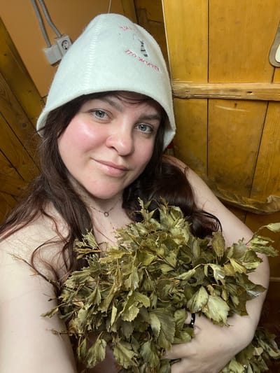 The image is a close-up selfie of a woman relaxing in a traditional Russian banya. The setting is an indoor space with warm, light-colored wooden plank walls and what appears to be a wooden ceiling, characteristic of a sauna or bathhouse. Electrical outlets and conduit wiring are visible on the wooden wall in the lower left.
The subject is a woman with long, dark brown hair and light-colored eyes. Her face shows a relaxed, content expression, with a slight smile and visibly flushed cheeks, suggesting she is in a hot environment. She wears a delicate gold necklace with a small circular pendant.
On her head, she wears a white felt sauna hat, which features red embroidered text. Clearly visible on the hat is the Russian phrase "ПО ЖИЗНИ" (PO ZHIZNI), which translates to "FOR LIFE" or "THROUGHOUT LIFE."
She is holding a large, verdant bundle of dried leaves, known as a "venik," commonly used in Russian banyas for whisking the body. The leaves appear to be from birch or oak. A dark-colored ring is partially visible on one of her fingers, partially obscured by the leaves. The overall scene depicts a moment of relaxation and cultural practice within a banya, located in Protasy, Russia.