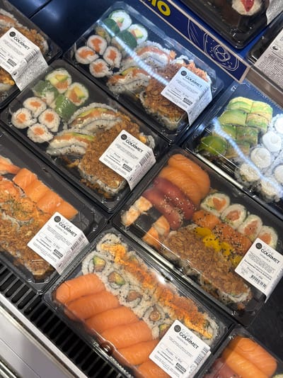 The image shows an arrangement of several containers of sushi on a shelf, likely in a refrigerated display at a store or market in Chianchitta, Italy. The containers are labeled with "SUSHI GOURMET" branding, along with details such as the type of sushi (e.g., "SNACKING BOX," "SPRING BOX," "HAISEN BOX"), price in euros (€), the date of packaging (29/05/2026), and the date by which it should be consumed (30/05/2026).
The types of sushi visible include rolls with various fillings (salmon, avocado, cream cheese, tuna), nigiri with salmon and shrimp, and variations with toppings like fried onions and spicy mayonnaise. Each container is transparent, allowing the sushi contents to be seen.
The containers are arranged in rows on a gray slatted shelf. The lighting suggests a retail environment, and the shelf construction suggests a refrigerated unit.