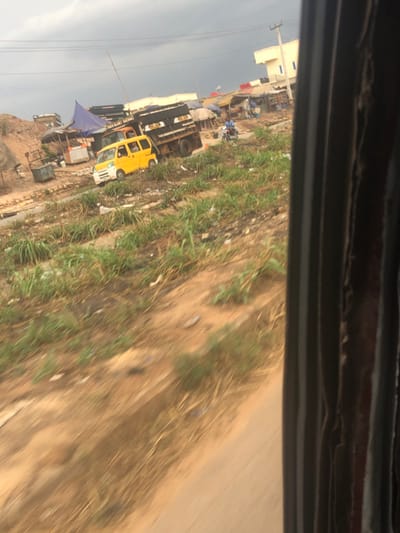 The scene depicts an unpaved urban or semi-urban area in Nwofia, Nigeria, viewed from the interior of a moving vehicle, indicated by the dark frame on the right and motion blur in the foreground. The immediate foreground consists of blurred brown dirt. Above this, an uneven dirt slope is covered with sporadic patches of green grass and scattered refuse, including plastic and other debris.
In the mid-ground, a yellow mini-van with a white front bumper is parked on this dirt incline, positioned ahead of a dark-colored flatbed truck. To the left of the yellow mini-van, a metal waste container is visible. Behind these vehicles and further up the slope, a series of informal market stalls are present, constructed from various materials including wood, corrugated metal, and blue tarpaulins.
One individual, wearing a blue shirt, is seated on a motorcycle positioned in front of these market stalls. Several other indistinct human figures are vaguely visible within the market area. In the background, beyond the market structures, a light-colored, multi-story building with a flat roof is discernible. Overhead power lines and utility poles are visible against an overcast, grey sky. No explicit actions or interactions among the individuals are clearly identifiable.