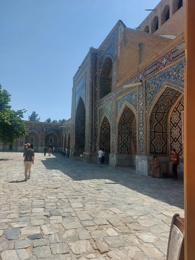 The image shows the Registan Square in Samarkand, Uzbekistan, a historical site known for its stunning madrasahs. The foreground features a cobblestone courtyard bathed in sunlight, casting sharp shadows. A man wearing a striped shirt and light-colored pants walks away from the camera towards a large, ornately decorated building. The building's facade is adorned with intricate blue, yellow, and white geometric tilework, set against a backdrop of brick. Arched doorways and windows punctuate the structure. In the middle ground, several other people are visible walking or standing, some near the building's entrance. To the left, a leafy green tree provides a contrast to the arid architecture. The sky is a clear, bright blue. The scene captures the grandeur and historical significance of the Registan Square.