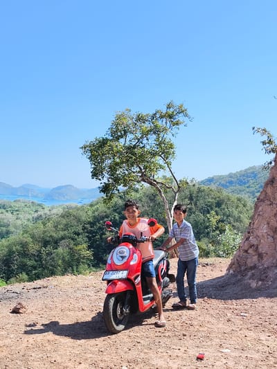 Two young boys are posing on and next to a red scooter on a dusty, elevated clearing overlooking a bay with islands and a tree with sparse foliage in the foreground. The boy on the scooter is wearing an orange t-shirt and blue shorts, while the other boy, standing to his right, wears a striped blue and white t-shirt and jeans. The scooter has a license plate that reads "EB 2297 G 11-29". The background shows lush green hills and mountains that slope down to a body of water with several boats. The sky is clear and blue. This scene is in Kaper, Indonesia, with its characteristic coastal and hilly terrain.