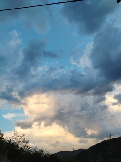 The image shows a cloudy sky above a landscape in Smilyan, Bulgaria. Dark clouds dominate the upper portion of the frame, contrasting with patches of blue sky. Lower in the image, clouds are illuminated with a golden hue, suggesting sunset or sunrise. In the foreground, the silhouette of trees and hills can be seen. A wire is present in the upper portion of the image.