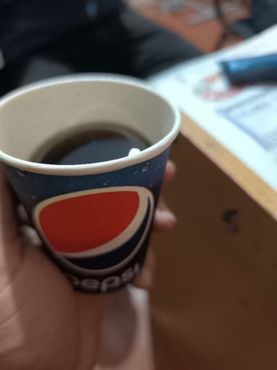 The scene depicts a close-up, eye-level view of a disposable paper cup containing a dark beverage, likely soda, held by a human hand in an indoor setting within Pakhtakor, Uzbekistan. The cup is primarily blue with water droplet condensation visible on its exterior and prominently features the red, white, and blue circular Pepsi logo. A small white object is present on the surface of the dark liquid inside the cup. A light-skinned hand, presumably a left hand, grips the lower section of the cup. In the blurred background, to the upper right, an open, light-colored book or document with visible text rests on a light brown surface, which appears to be a desk or table. A dark blue object, consistent with a pen or marker, lies partially on the open book. To the upper left, blurred dark fabric, likely part of clothing worn by an individual, is visible. The lighting is ambient and moderate. No distinct facial features or other individuals are clearly discernible.