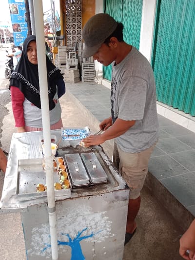 A street vendor cooks food on a metal cart decorated with a blue painted tree. He wears a gray t-shirt, khaki shorts, and a cap. He is standing to the right of the cart, using a tool to work on the food. A woman in a black hijab with a patterned edge and a pink shirt stands behind the cart, watching the vendor. Green metal doors are in the background.