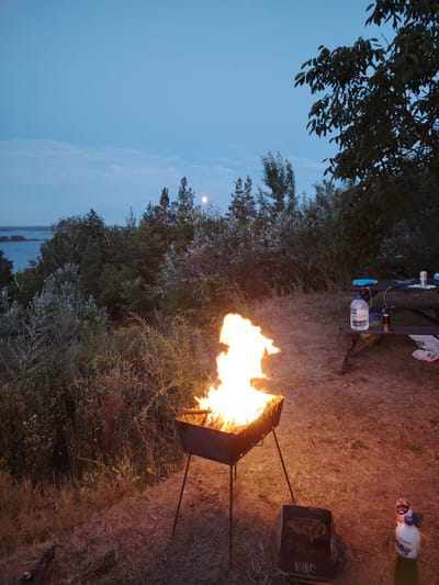 The image depicts an outdoor scene at dusk, likely in the city of Staiky, Ukraine. In the foreground, a portable charcoal grill with a bright fire is set up on dry grass. To the right of the grill, a cardboard box with Cyrillic text that appears to read "CAMPING" is visible, along with two plastic bottles, one of which has a purple cap and a label with Cyrillic text. Further to the right and slightly in the background, a wooden picnic table is set up. On the table, there are two plastic bottles of water, one larger and one smaller, a blue object, and a can. Behind the table, a dense cluster of trees and bushes creates a natural backdrop. In the distance, a body of water is visible, bordered by more trees and foliage. The sky above is a pale blue, with a faint haze, and a small, pale moon is discernible in the upper center of the frame. The overall impression is a campsite or picnic area during a twilight gathering.