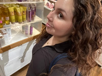 A woman with curly brown hair and a dark top stands next to a shelf stocked with Bath & Body Works products. The shelf has various lotions, perfumes, and sanitizers. The woman's face is turned towards the camera, and her eye is visible. There appears to be a shadow cast behind the shelf. The environment suggests the interior of a Bath & Body Works store.