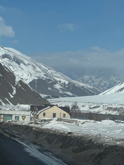 This image displays a snowy, mountainous landscape in Almasiani, Georgia, captured on a clear day with a blue sky and scattered clouds. In the immediate foreground, a dark, wet asphalt road is visible, bordered by high banks of dirty, compacted snow and slush to its right.
In the mid-ground, two buildings stand amidst deep snow. To the left, a light-colored, single-story structure with a flat roof presents green accents around its windows and a prominent black and yellow shield-shaped emblem on its exterior wall. Adjacent to it on the right is a two-story, cream-colored house featuring a dark, sloped roof and multiple windows. A small Georgian flag is affixed to the front of this house. Sparse, bare trees are scattered in the snowy terrain surrounding the buildings.
The background is dominated by expansive, snow-covered mountains and a wide valley. The mountain slopes to the left are steep and rugged, showing exposed rock alongside significant snow accumulation. The valley floor stretches into the distance, completely covered in white snow, with a visible road or pathway traversing it where faint, dark objects, possibly vehicles, are present. Farther in the background, additional snow-capped peaks rise, some partially obscured by a low band of clouds. Power lines are visible strung across the distant valley. No human figures are discernible within the scene.