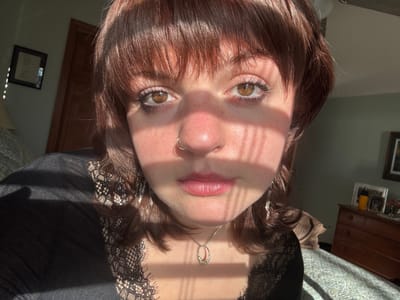 A woman with brown hair and eyes looks directly at the camera from a close-up, slightly tilted perspective. Sunlight streams across her face, creating shadows. She has a nose ring and is wearing a dark top with lace detailing around the neckline. A necklace is visible. The background includes parts of a room with furniture, like a dresser, and framed pictures on the wall.