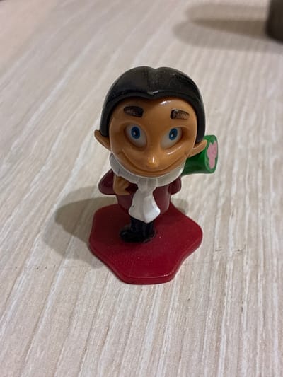A small, plastic figurine of a character with a dark, slicked-back hairstyle, blue eyes, and a wide smile stands on a red, hexagonal base. The figurine is wearing a dark jacket with a white scarf and appears to be holding a green object with pink accents in its right hand. The figurine is positioned on a light-colored, textured surface that resembles wood grain. The background is blurred, with shadows suggesting overhead lighting. There are no people, actions, or interactions depicted in the image. The location in the city of Melitopol, Ukraine, is not discernible from the provided image.
