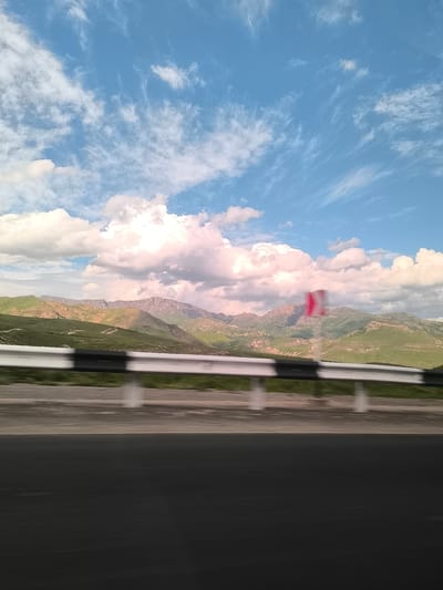 The image captures a vibrant mountainous landscape as seen from a moving vehicle, likely on a highway in Yelpin, Armenia. The scene is characterized by distinct horizontal layers.
In the immediate foreground, a dark asphalt road surface occupies the bottom third of the frame, exhibiting significant motion blur, which strongly suggests the photo was taken from a vehicle in transit. Just above the road runs a white and black striped metal guardrail, also blurred, reinforcing the sense of movement.
The midground features an extensive range of mountains and hills. These formations are largely covered in rich green vegetation, indicating a lush environment. Some higher, more distant peaks show lighter patches, possibly of exposed rock or lingering snow. The mountains are illuminated by bright daylight, casting subtle shadows and highlighting contours. A blurry red and white traffic sign, possibly a warning for a curve, is partially visible on the right side of the midground.
The sky dominates the upper portion of the image, displaying a clear, bright blue color. It is dotted with scattered white clouds of varying types. Fluffy cumulus clouds are prominent, some exhibiting a slight pinkish or purplish tint, possibly due to atmospheric conditions or the angle of the sunlight. Wispy, thin cirrus clouds are also visible, adding texture to the expansive sky.
The image conveys a sense of travel through a grand natural setting during a clear, sunny day. No people or discernible text are visible.