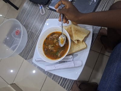 The scene is set indoors in the city of Close, Nigeria, at a table with a light grey and white striped surface. A dark-skinned individual, wearing blue denim trousers and brown slippers, is present. A white rectangular plate holds a white bowl of orange-brown soup containing meat chunks and green herbs. A metal spoon is submerged in the soup, held by the individual's right hand. Three slices of lightly toasted bread are also on the plate. Two sets of cutlery, each wrapped in a white napkin, are positioned below the bowl and toast. To the left of the plate, a translucent plastic bowl with a small red object inside is visible. A dark-colored menu or book, along with a mobile phone featuring a colorful beaded strap, are partially visible on the table in the background. The floor is light-colored tile with visible grout lines and reflections.