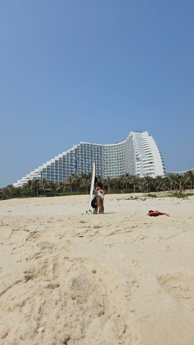 A high-angle, outdoor shot captures a sunny beach scene in Bac Cam Ranh, Vietnam. The image is oriented vertically, with a vast, clear blue sky dominating the upper half.

In the mid-ground, a sandy beach stretches across the frame. A woman, dressed in a white swimsuit or bikini, is standing or posing on the sand. She is positioned next to a white surfboard, which has subtle green and blue accents near its base. A small black object, possibly a bag, rests on the sand beside her. Further along the beach, to the right, an orange, somewhat deflated object is visible on the sand.

Behind the beach and the woman, a row of slender palm trees lines the boundary between the sand and developed land. Towering above the palm trees are large, modern white buildings, likely part of a resort or hotel complex. These buildings feature a distinctive curved architectural design with numerous rectangular windows, creating a striking backdrop. The structures are multi-storied, suggesting a significant presence.

The scene conveys a bright, clear day, characteristic of a tropical vacation destination. The lighting suggests mid-day or early afternoon with strong sunlight. The overall atmosphere is serene and leisurely, typical of a beach resort setting. There is no visible text in the image.