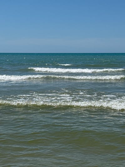 The image displays a seascape with clear blue skies on the left and the ocean on the right. Gentle waves are breaking, creating white foam along the shore. The water is a mix of deep blue and greenish hues, with sunlight reflecting off its surface, creating a shimmering effect. The scene is tranquil and serene, suggesting a peaceful day at the beach. The time of day appears to be daytime, with bright sunlight illuminating the water and sky. No people or objects are visible in the frame, focusing solely on the natural elements of the sea and sky. There is no visible text in the image. The location context suggests this scene is from Krasnyi, Russia.