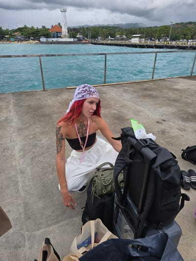 A red-haired woman wearing a white lace headscarf, black top, and white pants, with visible tattoos on her chest and left arm, stands on a gray concrete pier in Jungutbatu, Indonesia. She is surrounded by luggage, including a black suitcase and backpacks. The turquoise water of the sea is visible on the left, separated from the pier by a silver railing. In the background, the coastline is visible, with trees, buildings, and a white tower.