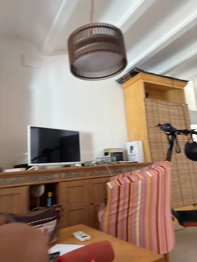 The image depicts an indoor scene, likely a living room, with furniture and an electric scooter. A television is mounted on a wooden cabinet. A striped chair is visible in the foreground, and a hanging light fixture is positioned on the ceiling. A tall, yellow cabinet with a bamboo screen is situated against the wall. An electric scooter with a black helmet is parked next to the cabinet. The room appears to be in an apartment or house, possibly in Benissa, Spain, judging by the presence of a white bag with a logo that might be from a local establishment. The overall lighting suggests daytime.