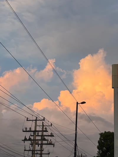 The image captures a dramatic sky over Managua, Nicaragua, filled with clouds illuminated by the setting sun. Power lines crisscross the frame, supported by utility poles, adding a sense of urban infrastructure against the natural beauty of the sky. A lone street light stands tall, its dark silhouette contrasting with the vibrant orange and pink hues of the clouds. In the background, the upper portion of a white structure with a red accent is visible on the right, possibly part of a building or sign. A dense green tree is partially visible in the lower right corner. The overall scene evokes a sense of twilight, blending the man-made elements with the atmospheric conditions of the Nicaraguan capital.
