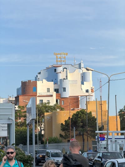 The image depicts a street scene in Chianchitta, Italy. The sky is light blue with faint clouds. On the left side of the image is a multi-story building with a varied facade: sections are pale blue and light beige, while others are brick red. The building has a sign that reads "Sporting Club Hotel". To the right of this building are yellow, weathered buildings. Several trees are interspersed between the buildings. Cars are parked along the street. Several people are visible including a man with a backpack, a man in a green shirt with sunglasses, and other people blurred in the background. Various street signs and shop signs are also visible.