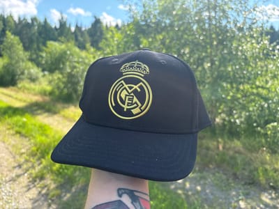 A person's arm is holding up a black baseball cap featuring the gold Real Madrid C.F. crest, which includes a crown and the letters "MCF." The background is a softly blurred, sunlit scene of greenery, likely trees and grass, with a clear blue sky with white clouds above. The sun appears to be high in the sky, suggesting it is daytime, and the lighting indicates fair weather. A dirt path or road is visible to the left of the cap. The person's arm has a tattoo. The overall impression is one of being outdoors in a natural, possibly wooded, setting.