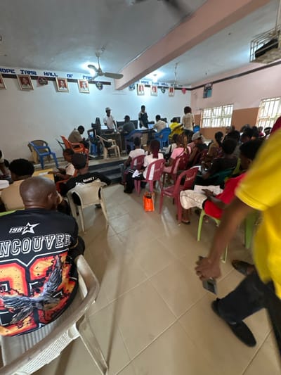 The scene is an indoor hall, identified by a wall banner as 'HOPE JAMB HALL OF FAME', located in Iriebe, Nigeria. Approximately 30-40 individuals, predominantly young adults, are present. The room features white walls, a light-tiled floor, a white ceiling with two operational fans, and windows with horizontal bars on the right. Framed portraits are displayed along the wall beneath the banner.

Individuals are seated on plastic chairs of various colors (pink, blue, green, white) arranged in rows, facing a frontal area. Many attendees are holding or reviewing papers; some forms display numbers such as '023' or '307'. An orange shopping bag is on the floor beside a pink chair.

At the front, a long wooden table serves as a workstation. Several individuals are positioned here; one male in a white cap and t-shirt stands behind the table. At least two laptops are visible on the table, operated by seated individuals.

In the foreground, a male subject is observed from behind, wearing a black t-shirt featuring '1987 VANCOUVER' and an eagle graphic. On the right, a subject in a yellow t-shirt and dark pants is partially visible, holding a dark-colored smartphone, seemingly in transit.