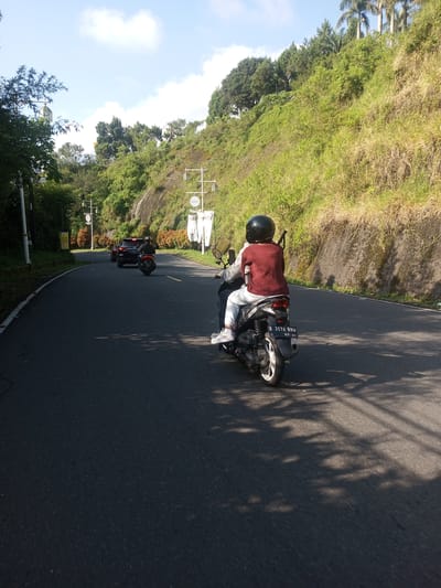 A road curves to the left through a mountainous area with lush greenery. In the foreground, a person is riding a scooter, wearing a helmet and a maroon sweater, away from the viewer. Behind them, another motorcycle and a dark-colored SUV follow the same path. Utility poles with signage are visible along the road's edge. Sunlight casts shadows on the asphalt. The location appears to be near a vegetated hillside with trees and bushes covering the slope. The setting suggests a rural or suburban area, not readily identified as Phouviang-Gnai, Laos.