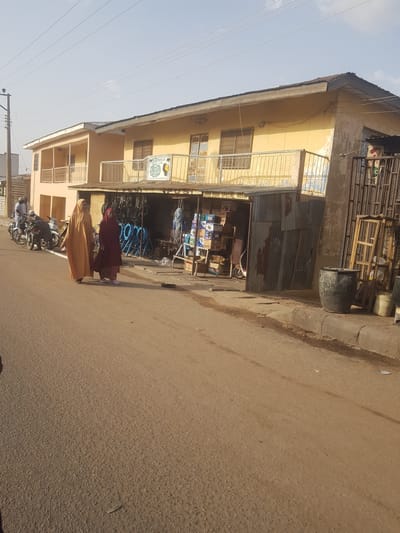 Two women in long dresses walk on the street in Dutsinma A, Nigeria. One woman is wearing a maroon dress, and the other is wearing a tan dress. A motorcycle is parked on the side of the street. Behind them is a two-story yellow building with a storefront, where goods are displayed. The sky is bright and clear.