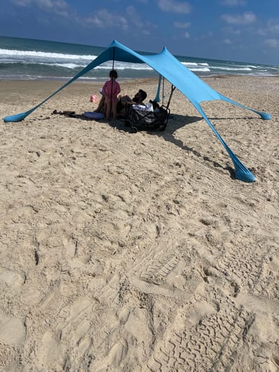 On the sandy beach of Gaash, Israel, a blue beach canopy provides shade for a family. A young girl in a pink rash guard stands under the canopy, facing away from the camera. Beside her, a man reclines on a mat, partially obscured by the canopy. A dark wagon with the word "Playa" is visible next to him. Scattered on the sand are a pink toy shovel, a towel, and a circular inflatable ring. The ocean with moderate waves is in the background, under a partially cloudy sky. The foreground is dominated by the textured sand of the beach, marked with footprints and tire tracks.
