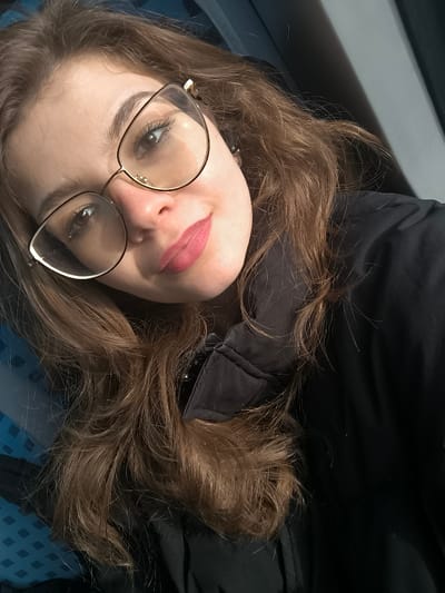 A close-up portrait depicts a single female subject with light brown, wavy hair, wearing gold-framed cat-eye eyeglasses. Her lips are colored with pink lipstick, and she exhibits a subtle smile while looking directly at the camera. She is dressed in a black jacket with a discernible collar texture. The background is indistinct, showing dark areas and a patterned blue and teal surface on the left. The specific location within the city of Kopanitsa, Bulgaria, is not visually identifiable from the image content. The scene is illuminated by strong directional light from the left, creating highlights on the subject's hair and glasses.
