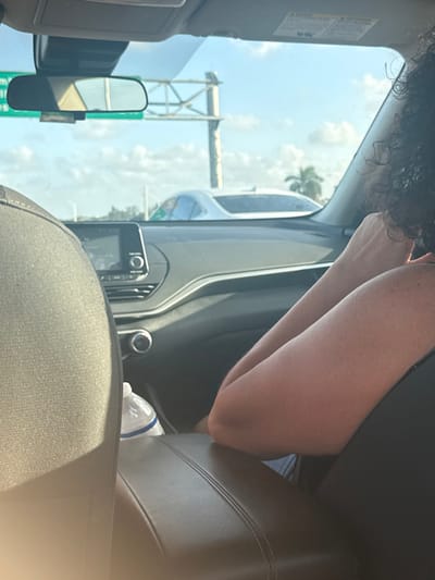 The image captures the interior front of a vehicle from a rear passenger's viewpoint, looking forward through the windshield onto a multi-lane highway. The setting is Hollywood, Florida, United States, identifiable by a green highway sign reflected in the rearview mirror, which clearly shows "Hollywood" and "27 Miles."
In the front passenger seat, an individual with dark, curly hair is visible from the upper back of their head and their right arm. Their right forearm rests on a dark, stitched center console/armrest. The vehicle's dashboard is dark, featuring a central infotainment screen and physical control buttons and knobs below it. Air vents are visible on the dashboard. A clear plastic water bottle with a blue label is positioned on the floor between the front seats. The top portion of the driver's seat headrest, upholstered in dark fabric, is visible on the left.
Outside the vehicle, the sky is clear and blue with scattered white cumulus clouds, indicating daylight. A large, grey metal highway gantry structure is prominent on the right side of the view through the windshield. A white sedan is visible in an adjacent lane to the right, driving parallel or slightly ahead. Other vehicles are faintly visible. In the background, palm trees and green vegetation line the highway, consistent with a subtropical environment. The scene depicts travel on a highway under clear, sunny conditions.