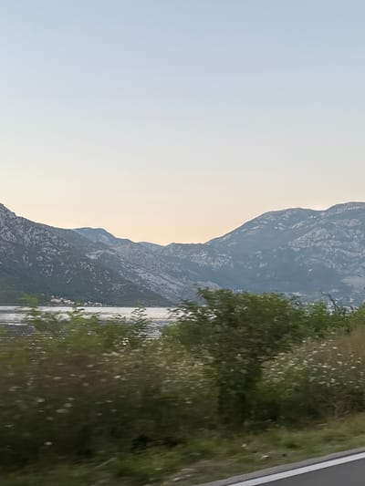 The image depicts a scenic view of the Bay of Kotor in Kostanjica, Montenegro. The foreground features dense green foliage, including trees and bushes with small white flowers, bordering a road with a white line. Beyond the vegetation lies the calm, reflective water of the bay, with ripples suggesting a gentle movement. In the mid-ground, a small coastal settlement with white buildings is visible nestled against the steep, mountainous terrain. The background is dominated by imposing, rocky mountains, characterized by their rugged textures and varying shades of gray and blue, suggesting a vast and dramatic landscape. The sky above is clear and pale, transitioning from a light blue at the top to a softer, warmer hue near the horizon, indicating either early morning or late evening light. The overall impression is one of natural beauty and tranquility, with the human settlement appearing small and integrated into the grand scale of the natural environment.
