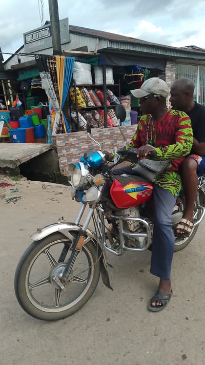 Two Black males are riding on a motorcycle in Agbarho, Nigeria. The motorcycle is red and silver and is in the foreground, facing right. The driver, wearing a colorful patterned shirt, a cap, and sunglasses, is holding the handlebars. The passenger, wearing a black shirt, is seated behind the driver. In the background, a store with various items for sale, including curtains and pillows, is visible. The environment appears to be an urban street with a cloudy sky.