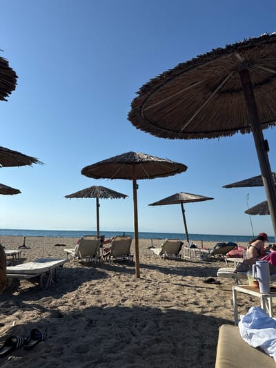 The image depicts a sandy beach on a clear day, likely in Angelochori, Greece. The foreground is dominated by sand and several thatched umbrellas providing shade. In the mid-ground, lounge chairs are arranged, some occupied by individuals. The sea is visible in the background, with a calm surface under a bright blue sky. A few people are present on the beach, some lounging on chairs and others near the water. The overall scene conveys a relaxed atmosphere typical of a seaside resort.