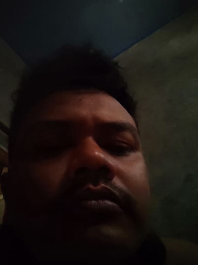 A close-up, low-angle selfie of a man's face is captured in a dimly lit room. The man appears to be of South Asian descent, with dark, curly hair that is styled upwards. His eyes are closed, and his face is illuminated by a light source from the front, casting shadows on the left side of his face. He has a prominent nose and a mustache above his upper lip. The background is dark, with what appears to be a textured wall on the right and a dark ceiling or upper wall on the left, suggesting an indoor environment. There is no discernible information about the specific location within Payakumbuh, Indonesia, in the image itself.