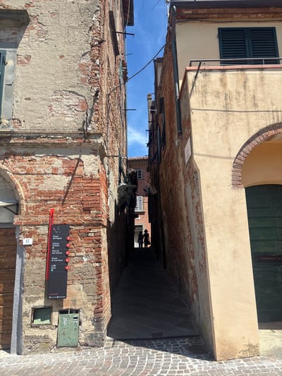 The image shows a narrow alleyway in the city of Peccioli, Italy. The alley is flanked by two buildings, one made of red brick and the other plastered in a light yellow hue. The brick building on the left displays signs indicating points of interest such as "Piazza del Popolo" and "Museo di Arte Sacra". There are two figures, silhouetted against the light at the end of the alley, walking away from the camera. The ground is paved with cobblestones. The sky is blue with scattered clouds.