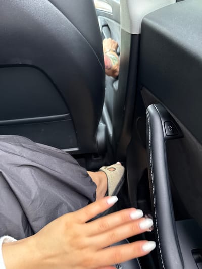 The image shows the interior of a vehicle, focusing on a person's feet and hand. A person wearing black pants and light-colored slip-on shoes is visible. The hand, with white painted nails, is in the foreground. In the background, another person with tattoos on their arm is visible reaching for a car door handle. The environment appears to be a car interior, with black leather seats and a door handle with a lock/unlock icon. The scene does not provide any information or context related to the city of Vahagnadzor, Armenia.