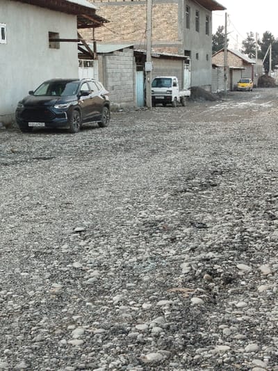 The image depicts a static, unpaved residential street scene in a city, identified as Andijan, Uzbekistan. The foreground consists of a wide expanse of rough, uneven gravel and small stones.
On the left side of the midground, a dark-colored SUV, identifiable by its front license plate "01 U 047 EB", is parked parallel to a light-colored, stucco-finished building. This building has a rectangular window and a smaller, higher opening, with a wooden beam extending from its side. Adjacent to this building is a grey cinder block wall, behind which an improvised wooden structure or roof is visible.
Further down the street, on the right side of the midground, a white compact flatbed truck is parked facing slightly towards the viewer. Behind it, a pile of dark, loose material, likely dirt or construction aggregate, is visible. Another cinder block wall and a two-story building with a brick upper section and concrete lower section, featuring multiple windows, are positioned to the right of the truck. A utility pole with overhead wires stands between the white truck and the two-story building.
The street continues into the background, where a yellow compact car is parked on the right side. Several other residential buildings line both sides of the road in the distance, predominantly single-story structures. The horizon features sparse trees and additional utility poles with wires. The sky is uniformly overcast, casting diffuse light over the scene. No individuals are present, and no actions or interactions are depicted. The environment suggests an urban residential area with unpaved roads and varying architectural styles.