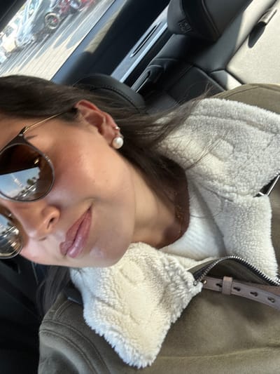 A selfie shows a woman with light skin, wearing round sunglasses and a pearl earring. She has dark hair. She's wearing a jacket with a white sherpa lining and a brown outer layer. The background appears to be the interior of a car, with a black leather seat visible. The scene is brightly lit, with reflections visible in the sunglasses lenses. The woman's face is angled towards the camera, and she is slightly smiling.