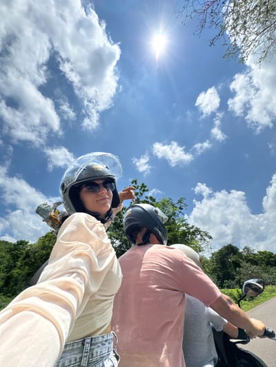 The image shows three individuals riding on a motorbike under a bright, sunny sky with scattered clouds. The person in the foreground, wearing a light-colored top and jeans, is looking towards the camera and wearing a helmet and sunglasses. To their right, another person is wearing a pink top and a helmet, and a third person, partially visible, is wearing a light gray shirt and a helmet. They are on a paved road surrounded by lush green trees and vegetation. The overall scene suggests a scenic journey on a motorbike, possibly in a rural or natural area. The specific location in Ham Thuan Bac, Vietnam, is not visually discernible from the image alone but could be inferred from metadata or external context.