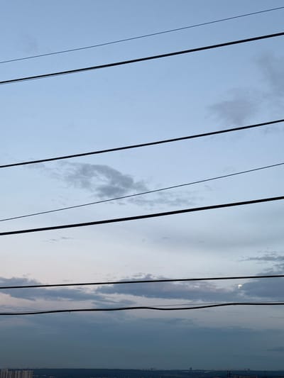 The image depicts a sky with scattered clouds and power lines stretching across the frame. The sun is partially visible through the clouds, suggesting it is either sunrise or sunset. The sky transitions from a light blue on the left to a darker, hazy blue on the right, where a distant city skyline is visible. The power lines are dark and angled, creating a linear pattern against the sky. The overall mood is serene and somewhat vast, with the focus on the expanse of the sky and the man-made elements that punctuate it. No people are visible in the image. The location is Otradnoye, Russia, which could explain the urban skyline in the distance. The time of day is likely early morning or late evening, given the lighting conditions and the presence of the sun partially obscured by clouds. The weather appears to be partly cloudy. There is no visible text in the image.