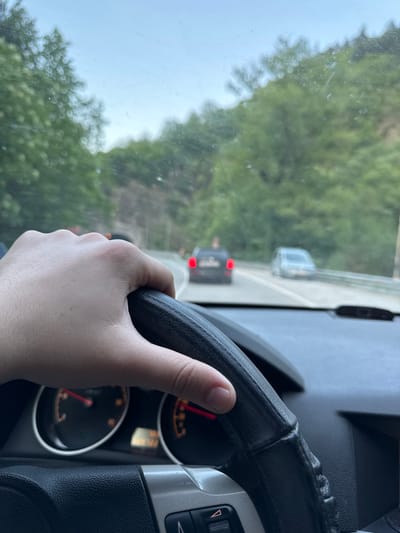 The image depicts the interior of a car, with a driver holding the steering wheel. Through the windshield, another vehicle can be seen on a road surrounded by trees. The speedometer is visible to the right of the steering wheel. The location is possibly near Бачковски манастир, Bulgaria, based on the surrounding environment.