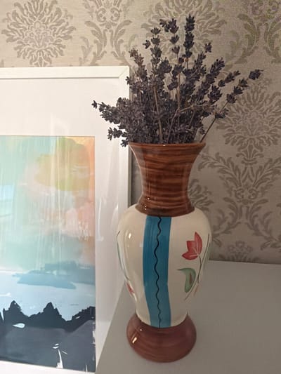 A close-up shot captures a decorative ceramic vase filled with dried lavender, placed on a white shelf against a backdrop of ornate, light-colored wallpaper. The vase is adorned with a pattern of blue stripes and red and green floral motifs. To the left of the vase, a portion of a framed abstract painting is visible, featuring soft colors like blue, yellow, and pink, and a dark, silhouetted landscape at the bottom. The scene appears to be an indoor setting, likely a home or a studio, given the decorative elements. The image is well-lit, with soft lighting that highlights the textures of the wallpaper and the vase. The overall impression is one of rustic elegance and artistic display. There is no indication of the specific location within Самахвалавіцкі сельскі Савет, Belarus, beyond it being an indoor environment.