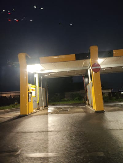 The image depicts an exterior scene at night in Laagri alevik, Estonia, featuring a brightly lit car wash facility. Two yellow support pillars, adorned with black and yellow striped bands near their tops, frame the entrance to the car wash bays. The pillars have signage and what appear to be payment or control terminals attached to them. One pillar also displays a red circular "no entry" sign with a horizontal white bar.
The car wash structure itself has a white canopy with yellow accents, illuminated by overhead lights, casting strong reflections on the wet asphalt ground. The ground appears damp and possibly recently rained upon, with visible streaks and patches of water. Beyond the car wash structure, the background is dark, with some distant lights and faint foliage suggesting an outdoor, possibly semi-rural or industrial area on the outskirts of the town. The overall lighting is artificial and dramatic, highlighting the structure against the night sky, which is mostly dark with a few faint, distant lights visible at the top of the frame. There are no people or vehicles clearly visible within the immediate vicinity of the car wash.