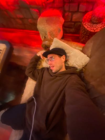 A young man is casually lounging indoors, bathed in dramatic red lighting. He lies on his side, wearing a dark brown hooded sweatshirt with a visible white drawstring, a dark baseball cap, and clear-rimmed glasses. His head rests on a light-colored, fluffy pillow, and he looks towards the viewer with a relaxed, slightly smiling expression.

The man is positioned on a dark-colored upholstered surface, likely a sofa or bed. To his left, a prominent, light-colored decorative object resembling a stylized human head or a large vase with intricate textures is visible. Behind this object, a wall with a brick-like pattern is illuminated by the red light, adding to the ambient glow. A portion of another cylindrical, textured object is also visible in the upper left corner. In the bottom right, a small part of a keyboard or similar electronic device can be seen. The overall setting appears to be a dimly lit room, possibly a themed lounge area, with the red lighting creating a distinct, moody atmosphere. No visible text is present in the image. This scene is located in Moračko Trebaljevo, Montenegro.