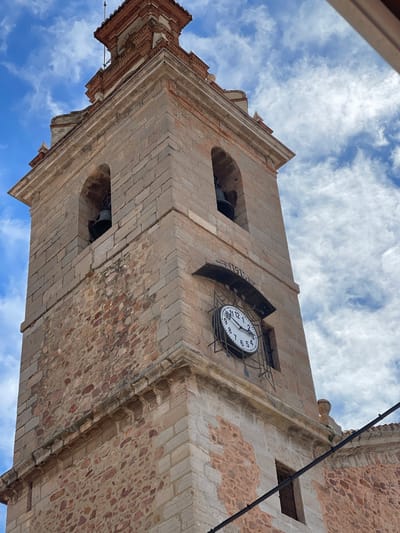 The image shows the bell tower of the Church of the Saints John in Puçol, Spain, from a low angle. The tower is made of stone and brick and has a clock on one side. There are visible bells in arched openings on the tower. The sky is cloudy.